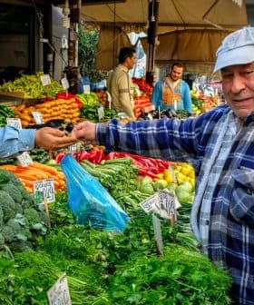 A man in a blue cap interacts with a vendor at a vegetable market filled with various fresh produce. - Olive Oil Times