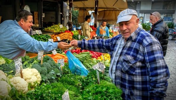 A man in a blue cap interacts with a vendor at a vegetable market filled with various fresh produce. - Olive Oil Times