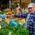 A man in a blue cap interacts with a vendor at a vegetable market filled with various fresh produce. - Olive Oil Times