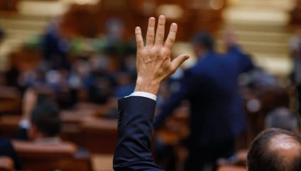 A hand raised in a formal setting, possibly during a vote in a legislative assembly. - Olive Oil Times