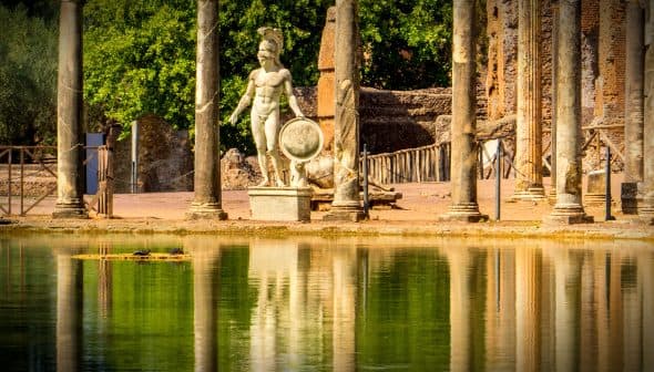 Statue of a Roman warrior holding a shield near ancient columns and water reflection. - Olive Oil Times
