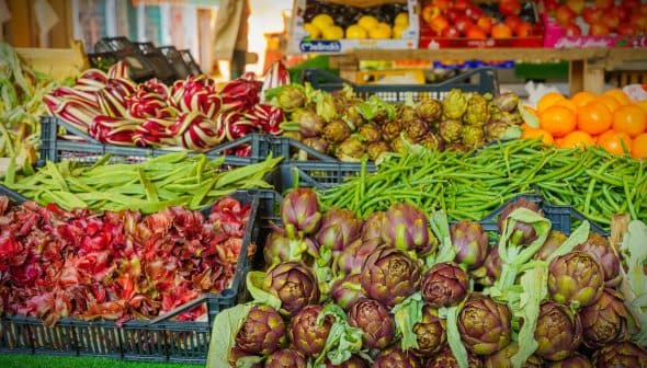 Display of assorted fresh vegetables and fruits including artichokes, green beans, and oranges at a market. - Olive Oil Times