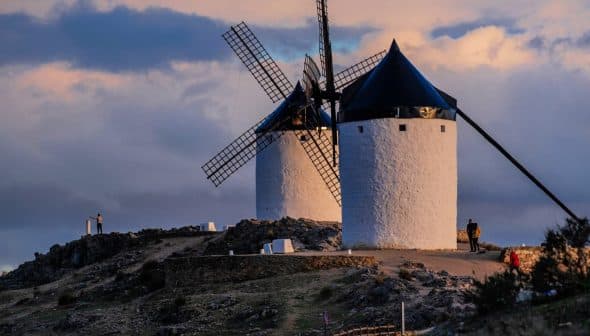Two traditional windmills with black blades and white stone structures situated on a hilltop. - Olive Oil Times