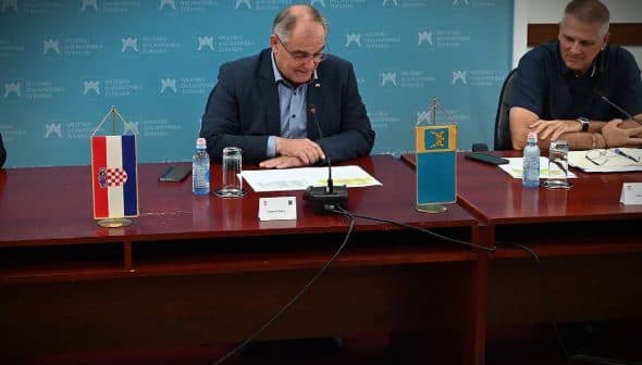 A person sitting at a table with flags of Croatia and another entity displayed on stands. - Olive Oil Times