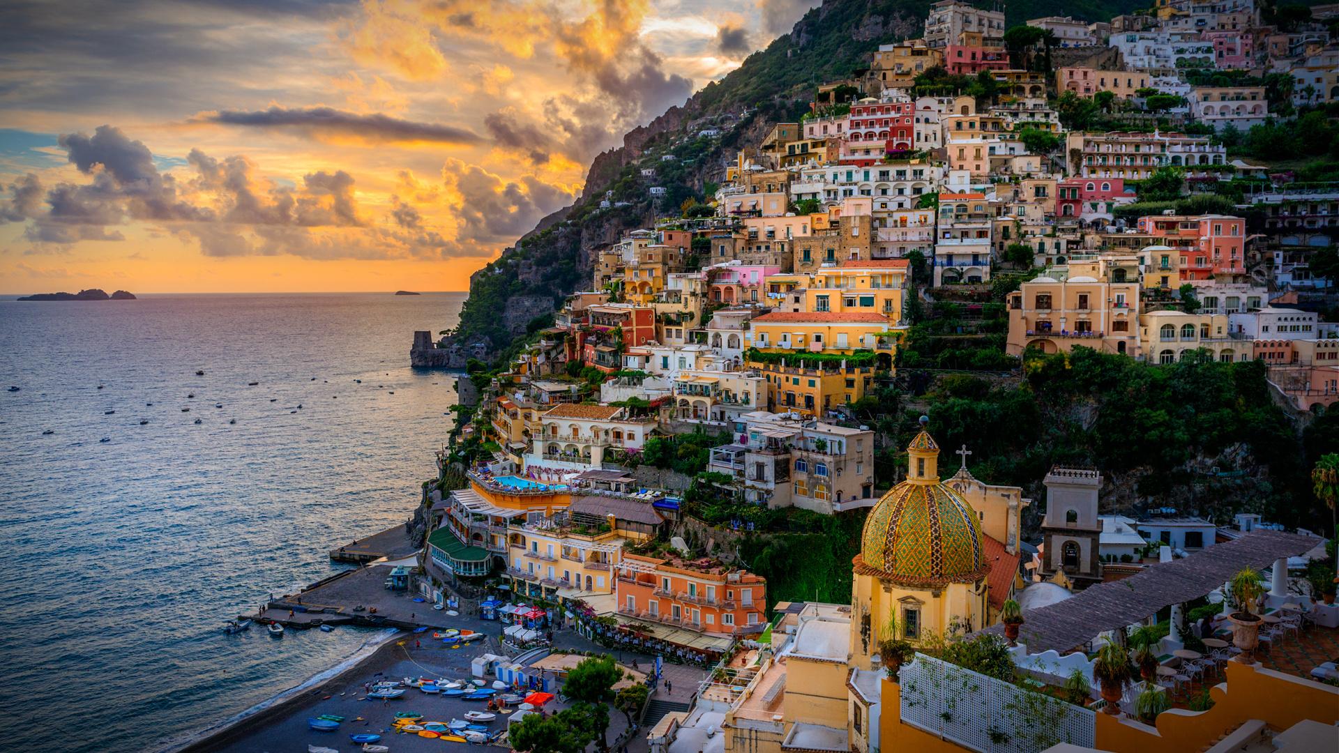 View of the coastal town of Positano with colorful buildings and a beach during sunset. - Olive Oil Times
