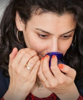 Woman with curly hair smelling from a blue glass container in a neutral setting. - Olive Oil Times