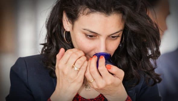Woman with curly hair smelling from a blue glass container in a neutral setting. - Olive Oil Times