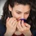 Woman with curly hair smelling from a blue glass container in a neutral setting. - Olive Oil Times