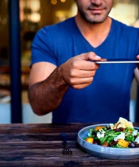Individual holding a smartphone to take a photo of a salad dish on a wooden table. - Olive Oil Times