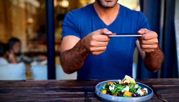 Individual holding a smartphone to take a photo of a salad dish on a wooden table. - Olive Oil Times