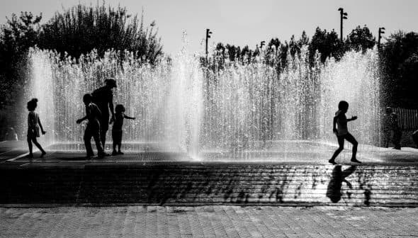 Silhouettes of children playing in a water fountain with water spraying in the background. - Olive Oil Times