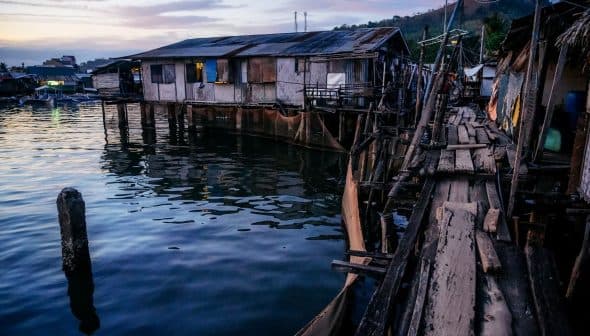 Wooden stilt houses built over water with a wooden walkway leading to them during twilight. - Olive Oil Times
