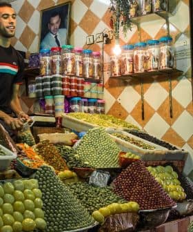 A market stall displaying a variety of olives and spices, with a vendor and a customer present. - Olive Oil Times
