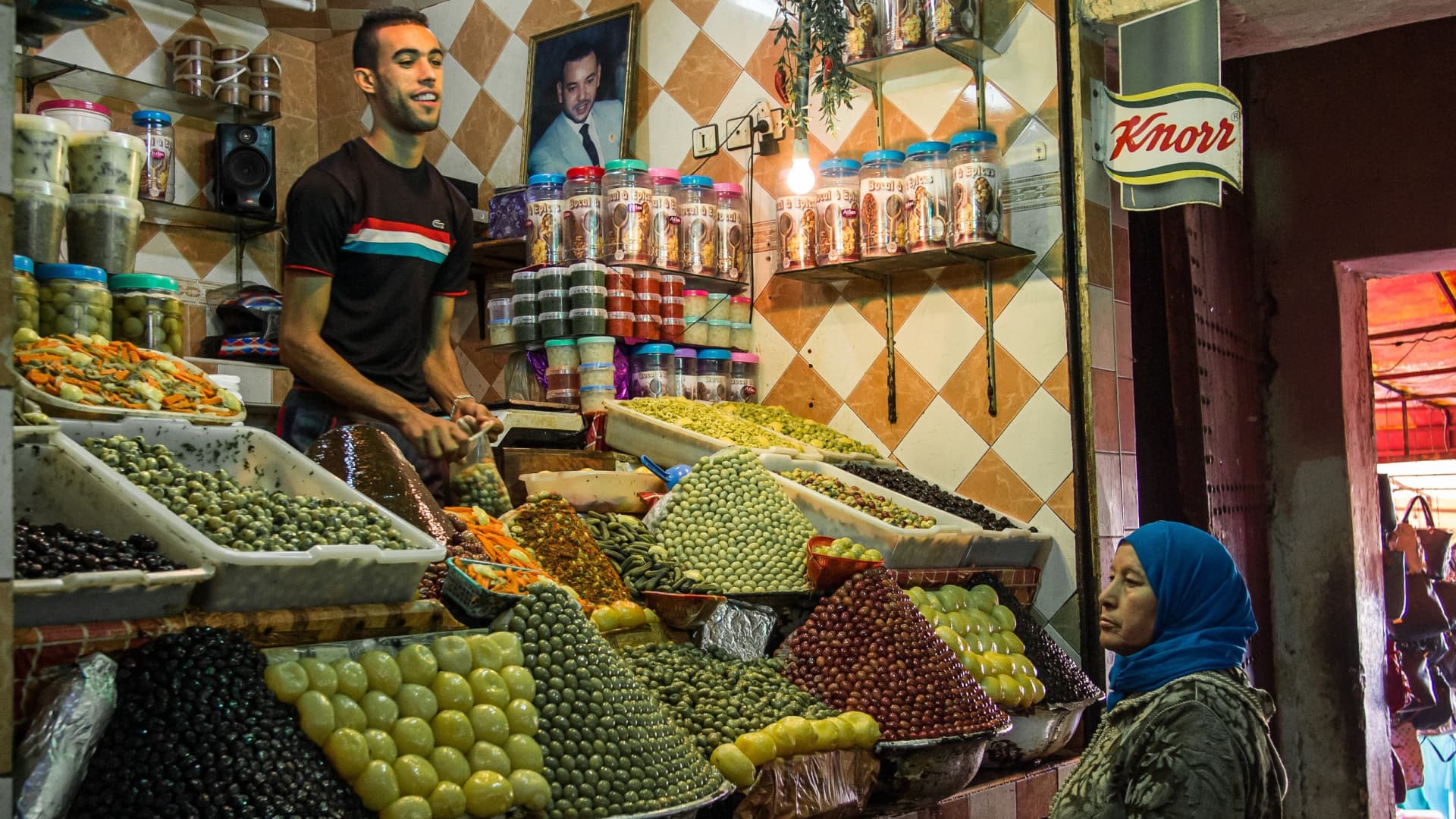 A market stall displaying a variety of olives and spices, with a vendor and a customer present. - Olive Oil Times