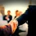 Two business professionals shaking hands in a formal setting with blurred figures in the background. - Olive Oil Times