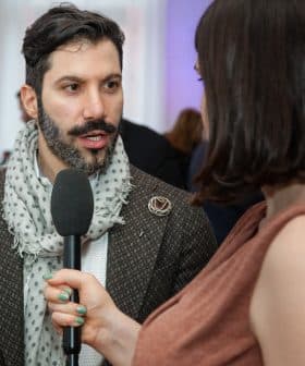Man with a beard wearing a patterned coat and scarf speaking into a microphone during an interview. - Olive Oil Times