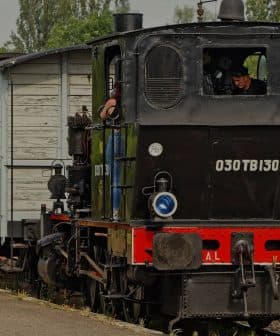A black steam locomotive with a blue and brown passenger carriage at a train station. - Olive Oil Times