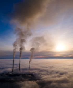 Three smoke stacks emitting smoke above a layer of clouds during sunset. - Olive Oil Times