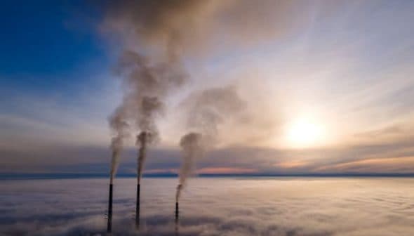 Three smoke stacks emitting smoke above a layer of clouds during sunset. - Olive Oil Times