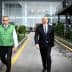 Three men walking on a pathway, two wearing green vests and one in a suit. - Olive Oil Times