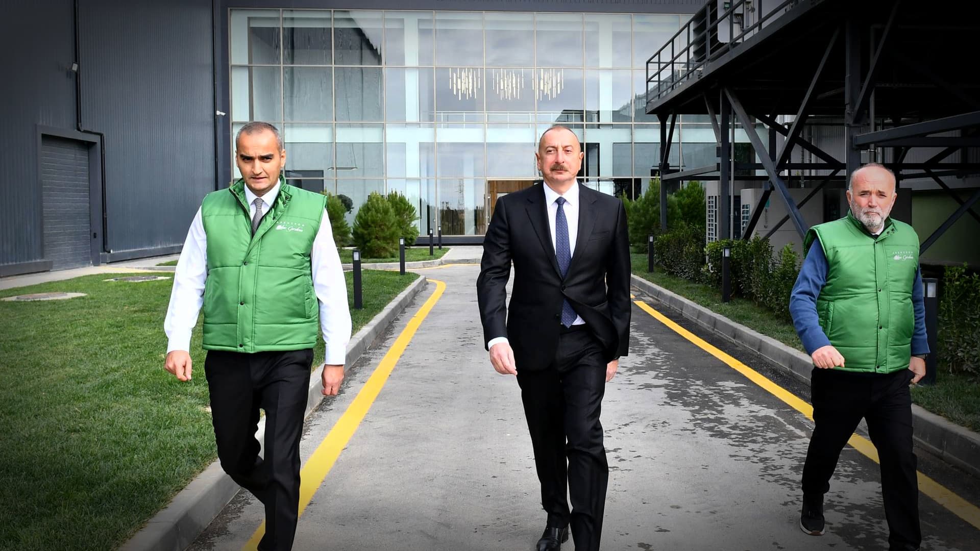 Three men walking on a pathway, two wearing green vests and one in a suit. - Olive Oil Times
