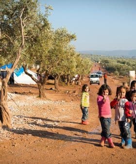 Group of children standing in an olive grove with tents in the background. - Olive Oil Times