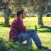 A man in a red plaid shirt sitting on a rock under an olive tree, holding a bottle. - Olive Oil Times
