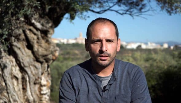 A man sitting in front of a large olive tree, looking directly at the camera with a serious expression. - Olive Oil Times