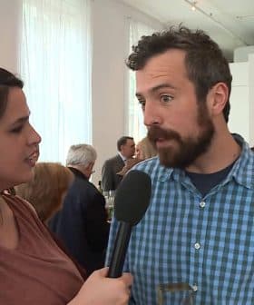 A man in a blue checkered shirt speaking to a woman holding a microphone at an event. - Olive Oil Times