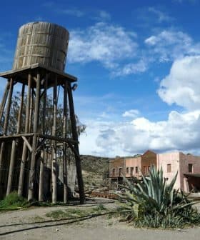 A wooden water tower stands beside a desert town with various buildings and a cactus. - Olive Oil Times