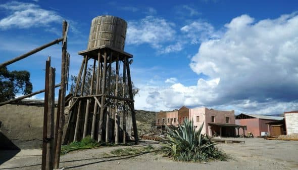 A wooden water tower stands beside a desert town with various buildings and a cactus. - Olive Oil Times