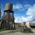 A wooden water tower stands beside a desert town with various buildings and a cactus. - Olive Oil Times