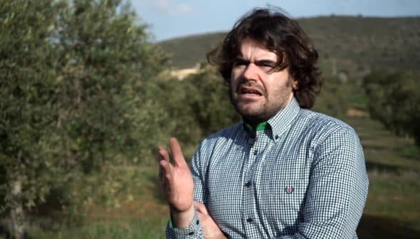 Man wearing a checkered shirt gesturing while standing in an olive grove. - Olive Oil Times