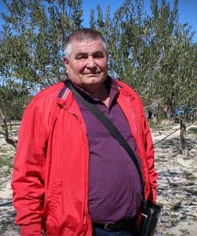 Man wearing a red jacket and purple shirt standing in an olive grove with trees in the background. - Olive Oil Times