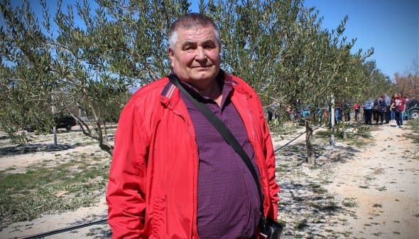 Man wearing a red jacket and purple shirt standing in an olive grove with trees in the background. - Olive Oil Times