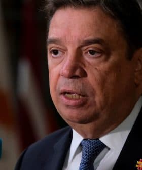 A man in a suit speaking at a press conference with flags in the background. - Olive Oil Times