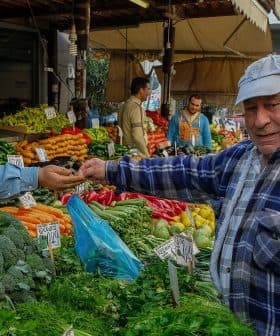 Man in a blue plaid shirt interacting with a vendor at a vegetable market stall. - Olive Oil Times