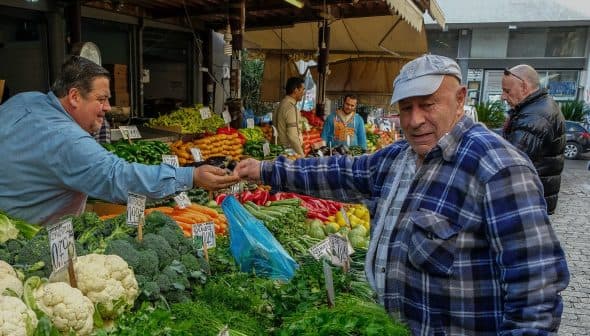 Man in a blue plaid shirt interacting with a vendor at a vegetable market stall. - Olive Oil Times
