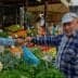 Man in a blue plaid shirt interacting with a vendor at a vegetable market stall. - Olive Oil Times