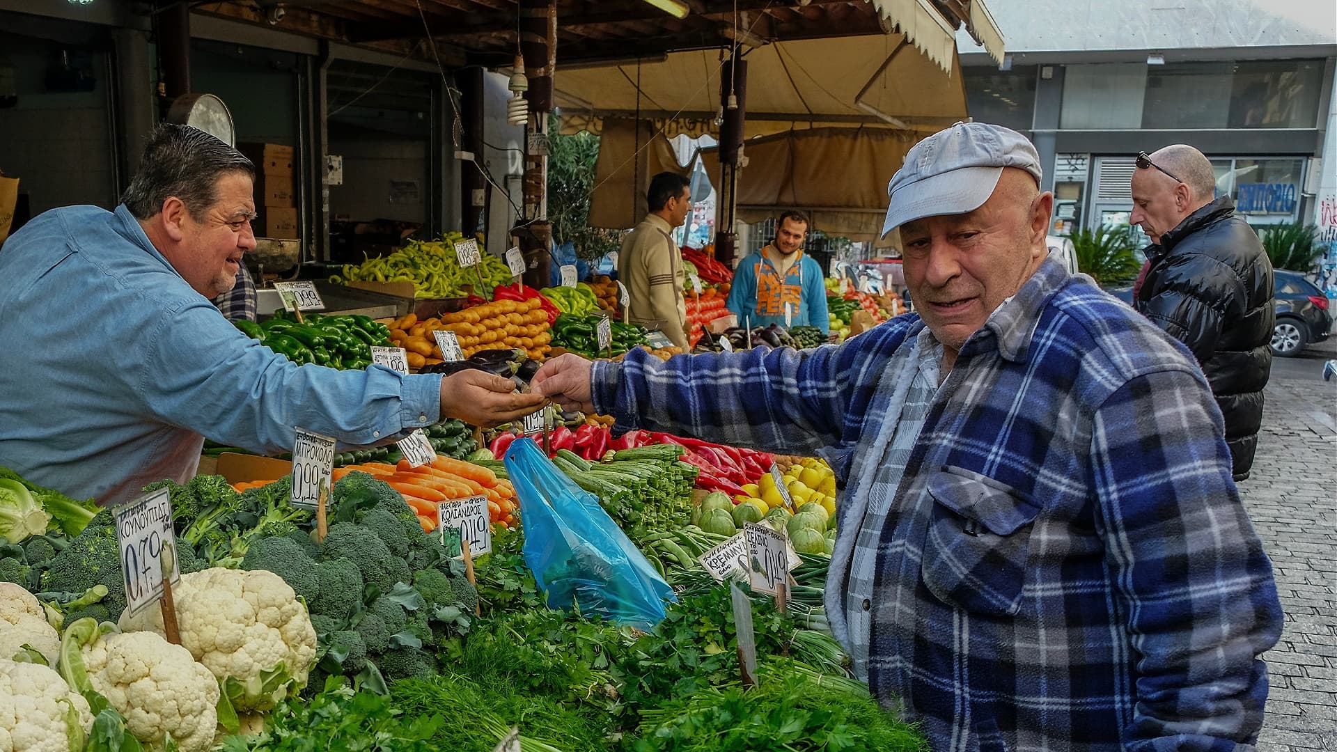 Man in a blue plaid shirt interacting with a vendor at a vegetable market stall. - Olive Oil Times