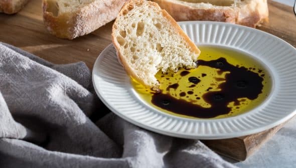 Slice of bread dipped in olive oil with balsamic vinegar on a white plate. - Olive Oil Times