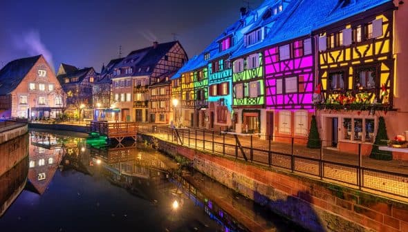 Row of half-timbered houses illuminated in various colors along a canal at night. - Olive Oil Times