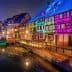 Row of half-timbered houses illuminated in various colors along a canal at night. - Olive Oil Times