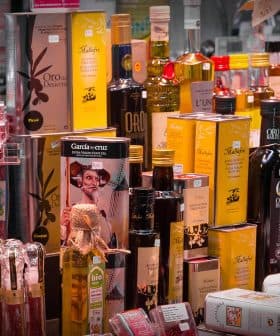 Various bottles and jars of olive oil and condiments displayed on a shelf in a market. - Olive Oil Times