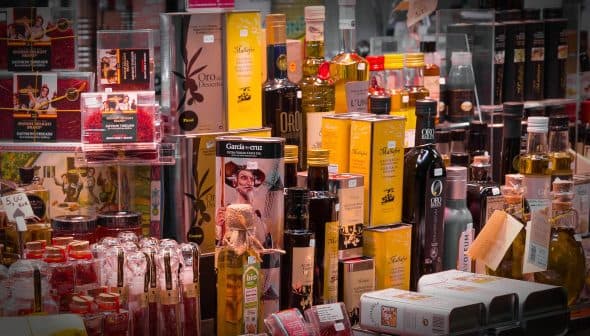 Various bottles and jars of olive oil and condiments displayed on a shelf in a market. - Olive Oil Times