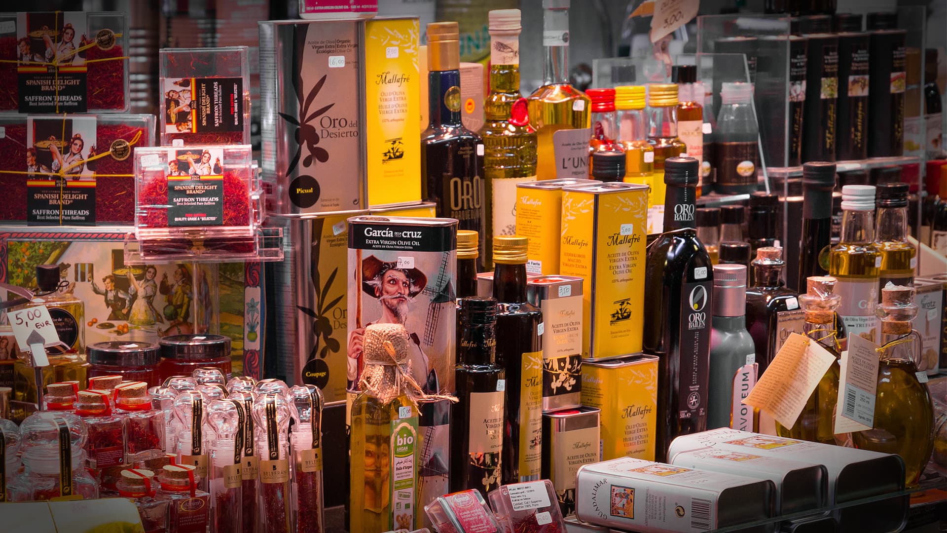 Various bottles and jars of olive oil and condiments displayed on a shelf in a market. - Olive Oil Times