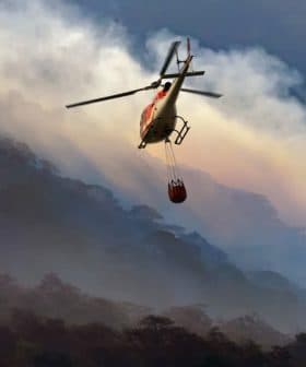 Helicopter flying over a mountainous area with a water bucket suspended below it. - Olive Oil Times
