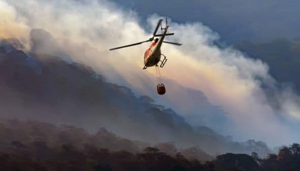 Helicopter flying over a mountainous area with a water bucket suspended below it. - Olive Oil Times