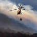 Helicopter flying over a mountainous area with a water bucket suspended below it. - Olive Oil Times