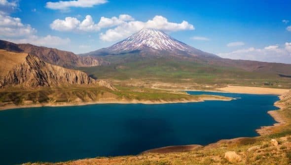 Snow-capped mountain with a clear blue sky above a large lake surrounded by hills. - Olive Oil Times